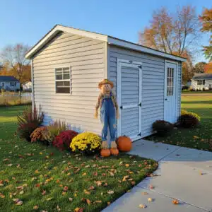Slightly used 10x14 vinyl A-frame shed in stone gray with charcoal gray trim, featuring a 9-lite door, standard door, window, fall flowers, and a scarecrow.