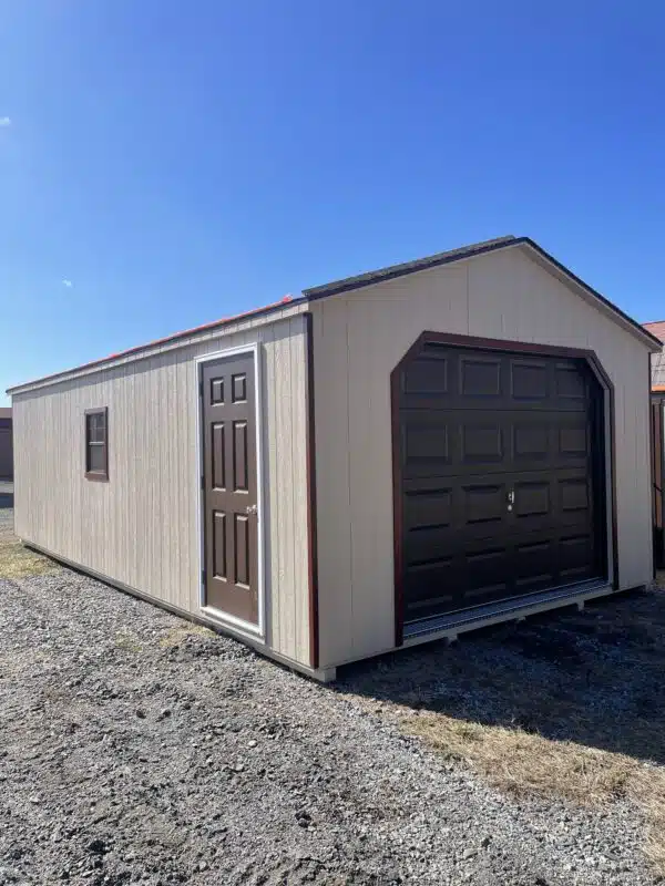Tan A-frame garage with brown overhead door, brown walk-in door, redwood trim, and two brown-trimmed windows, set on a gravel lot under a clear blue sky.