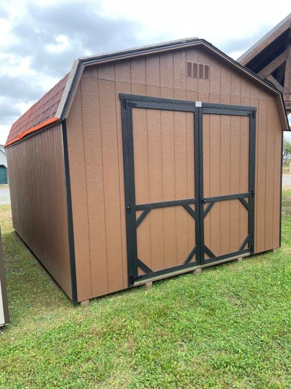 Chestnut brown Dutch barn-style storage shed with black double doors and trim, set on grass under a cloudy sky.