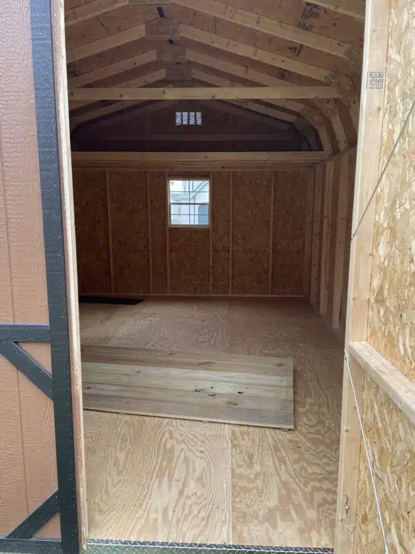 Unfinished interior of a Dutch barn-style shed with exposed studs, plywood floor, loft area, and a window at the far end.