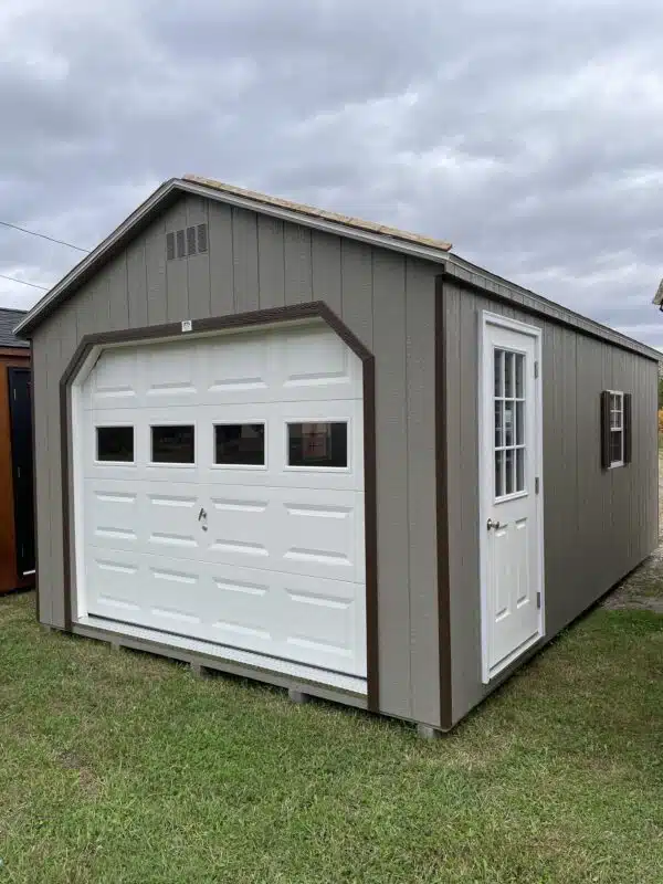 Gray A-frame single-car garage with white overhead door featuring windows, white entry door with glass, and brown trim, set on grass under a cloudy sky.
