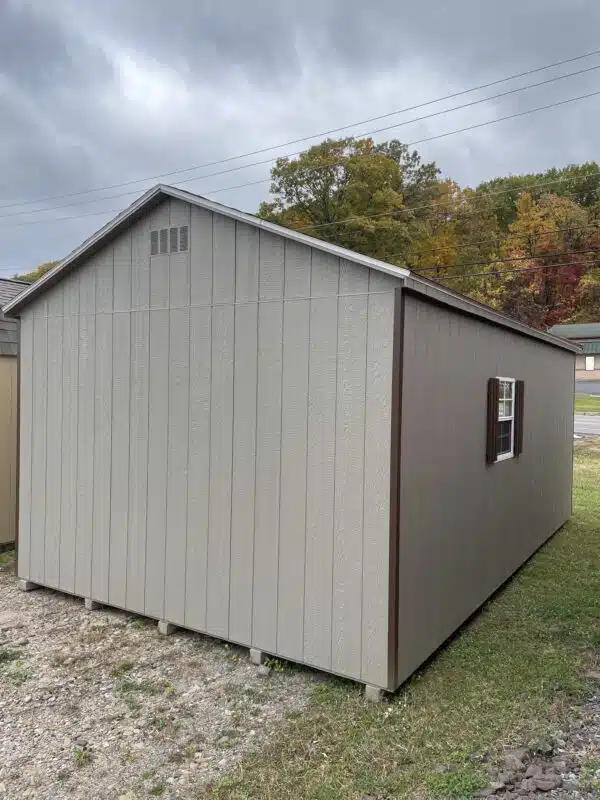 GP Clay Smartside A-frame garage with brown trim, driftwood roof, white 9x7 overhead door with windows, side entry door, window with shutters, and ramp, set on grass under a cloudy sky.