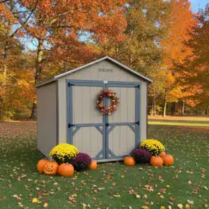 Gap gray Carolina Cottage shed with blue gray double doors and trim, decorated with a fall wreath, pumpkins, and mums, set on green grass with colorful autumn trees in the background.