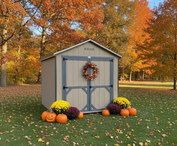 Gap gray Carolina Cottage shed with blue gray double doors and trim, decorated with a fall wreath, pumpkins, and mums, set on green grass with colorful autumn trees in the background.