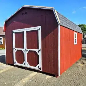 Red barn-style storage shed with white double doors and trim, gambrel roof, and a single window on the side, set on a gravel lot under a blue sky.