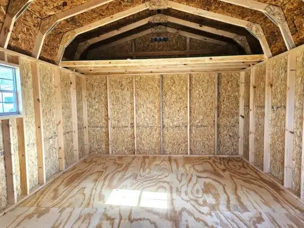 Unfinished interior of a Dutch barn-style storage shed with exposed studs, plywood floor, and a loft area under a gambrel roof.