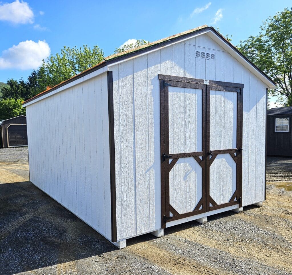 White A-frame storage shed with dark brown double doors and trim, shown from a front corner angle on a gravel lot under a blue sky.
