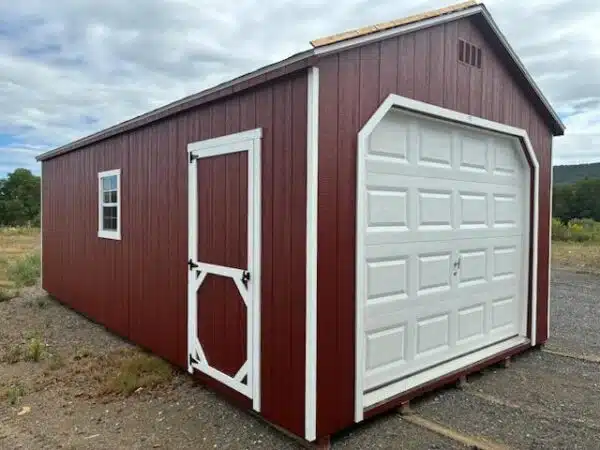 Red A-frame garage shed with white overhead garage door, white walk-in door, and white-trimmed window, set on a gravel lot.