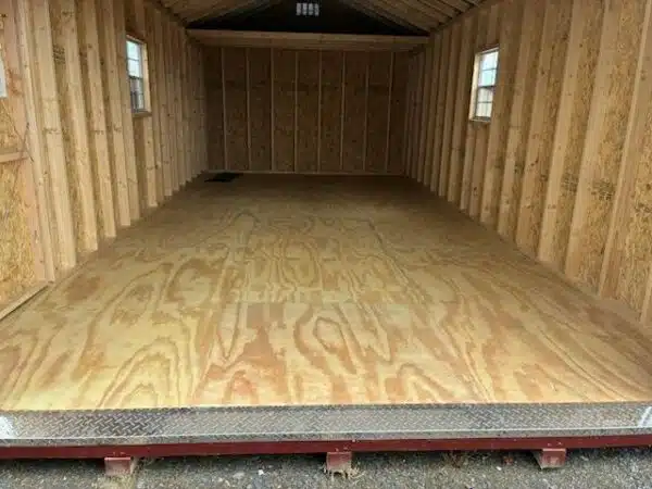 Unfinished interior of a 12x24 A-frame garage with exposed studs, plywood flooring, two side windows, and a metal tread plate at the entrance.
