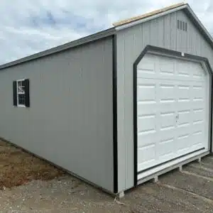 Gray single-car garage shed with a white overhead door, black trim, and a side window with black shutters, set on a gravel lot.