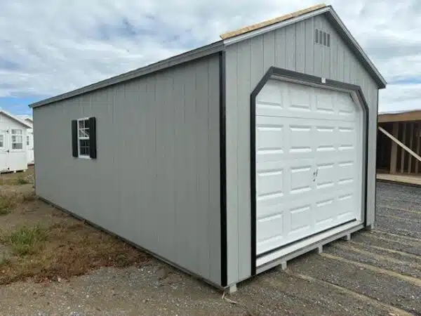 Gray single-car garage shed with a white overhead door, black trim, and a side window with black shutters, set on a gravel lot.