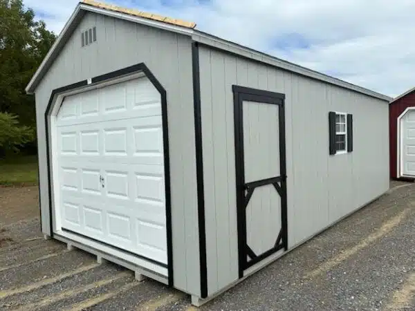 Gray A-frame garage shed with a white overhead door, black trim, black side entry door, and a side window with black shutters, set on a gravel lot.