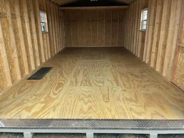 Unfinished interior of a garage shed with exposed studs, plywood flooring, two side windows, and a metal tread plate at the entrance.