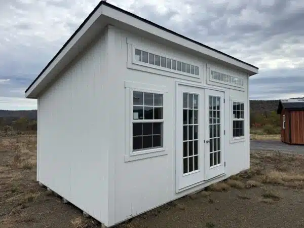 White leantoo office shed with double glass doors, transom windows, and three large windows, set on a gravel lot under a cloudy sky.