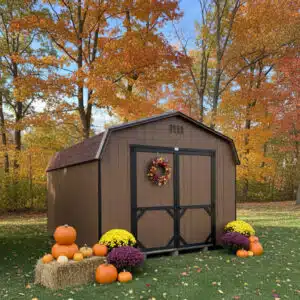 Chestnut brown Dutch barn-style shed with black trim, decorated with a fall wreath, pumpkins, mums, and hay bale, set against colorful autumn trees.
