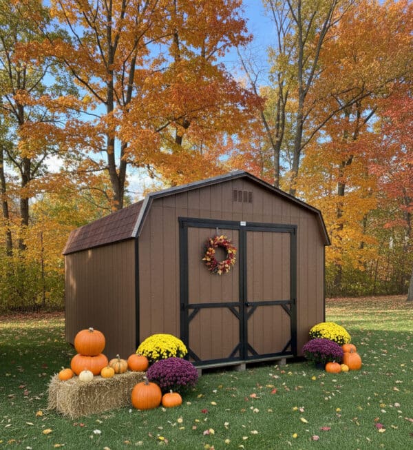 Chestnut brown Dutch barn-style shed with black trim, decorated with a fall wreath, pumpkins, mums, and hay bale, set against colorful autumn trees.