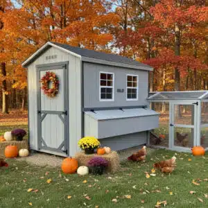 Gray 6x8 chicken coop with dark gray trim, exterior nesting box, attached run, and fall wreath, surrounded by pumpkins, mums, hay bales, and chickens, set on grass with colorful autumn trees.