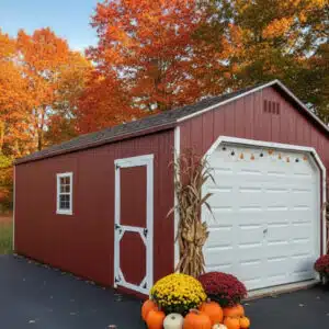 Red A-frame garage with white doors and trim, decorated with pumpkins, mums, and cornstalks for fall, set against colorful autumn trees.