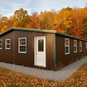 Brown 28x48 finished recreational cabin (Stock Model 1065U) surrounded by autumn trees, with gravel foundation and white entry door.