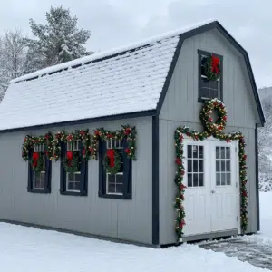 Gray two-story shed decorated for Christmas with wreaths, garland, and red bows, surrounded by snow.