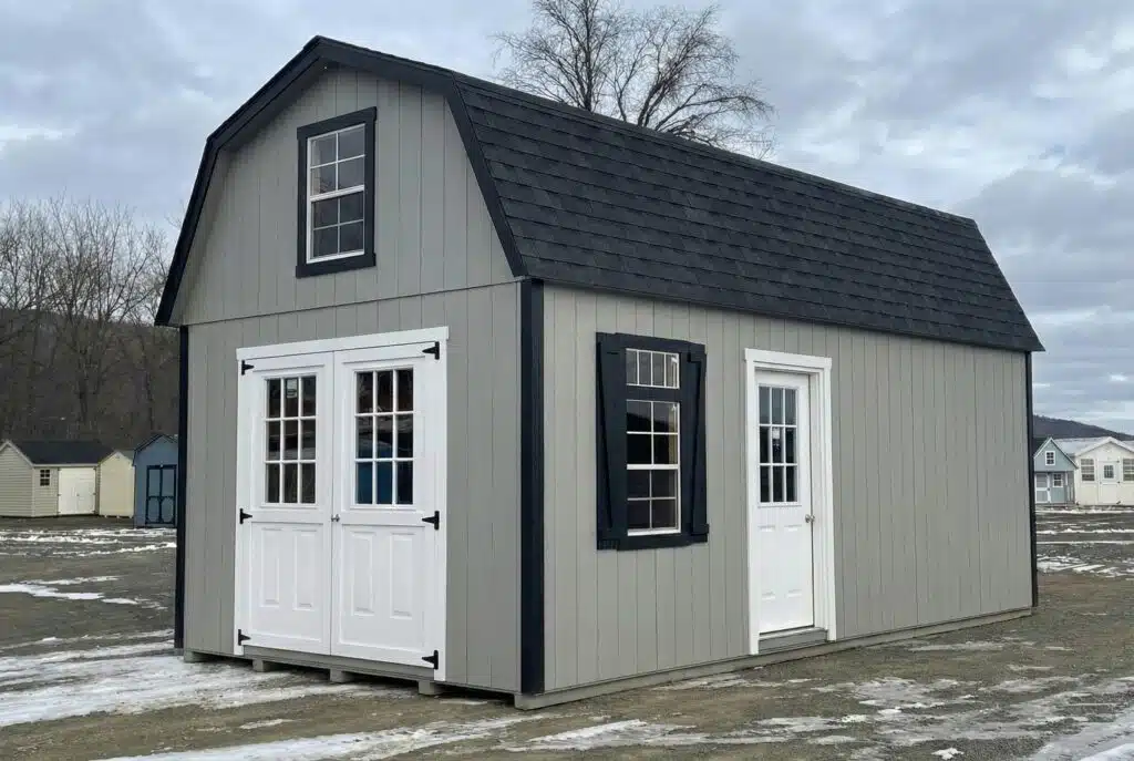 Gray two-story shed with black roof, white trim, and multiple windows, shown on a lot with other storage buildings in the background.