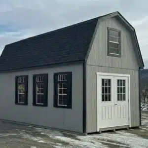 Two-story gray shed with black shutters and white double doors, set outdoors on a winter day.