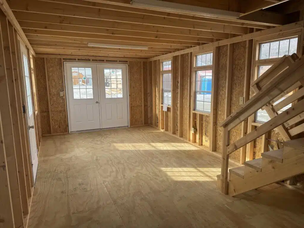 Bright unfinished interior of a two-story shed with wood floors, exposed framing, multiple windows, and stairs.