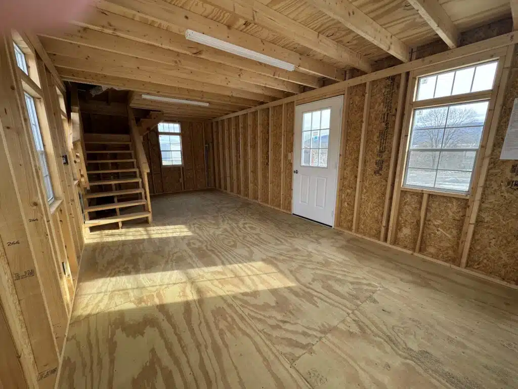 Interior view of an unfinished two-story shed with wood framing, windows, and a staircase leading to the upper level.