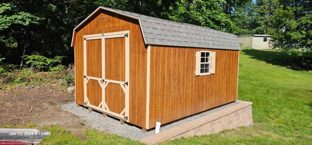 Brown Dutch barn-style storage shed with beige trim and window shutters, set on a gravel pad in a sunny, green backyard.