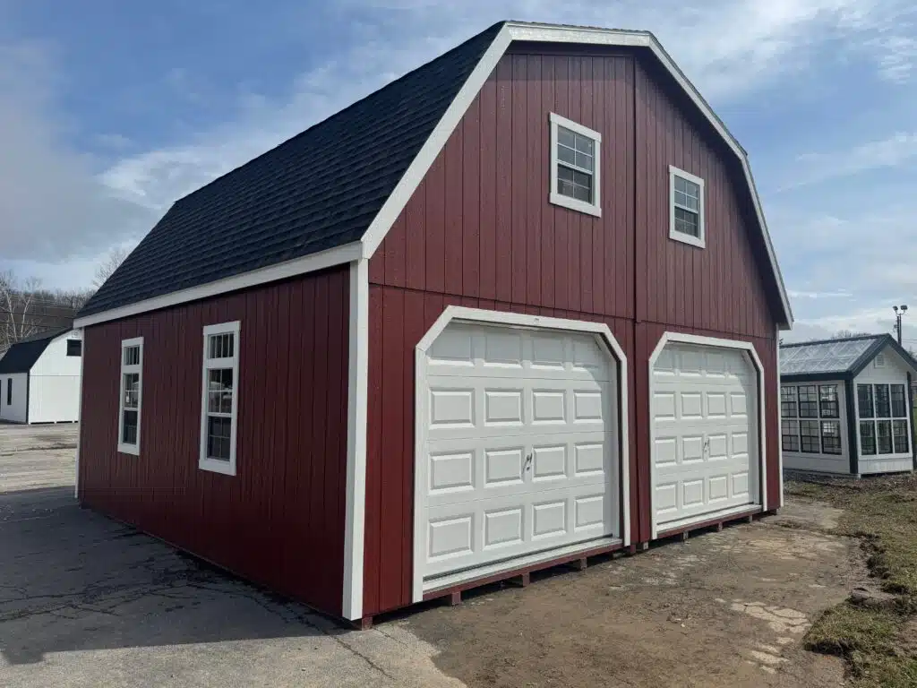 Red 24x24 two-story garage with white trim, black roof, two white garage doors, upper windows, side windows, and ridge vent shown from the front-left angle on a display lot