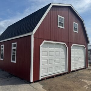 Red 24x24 two-story garage with white trim, black roof, two white garage doors, upper windows, side windows, and ridge vent shown from the front-left angle on a display lot