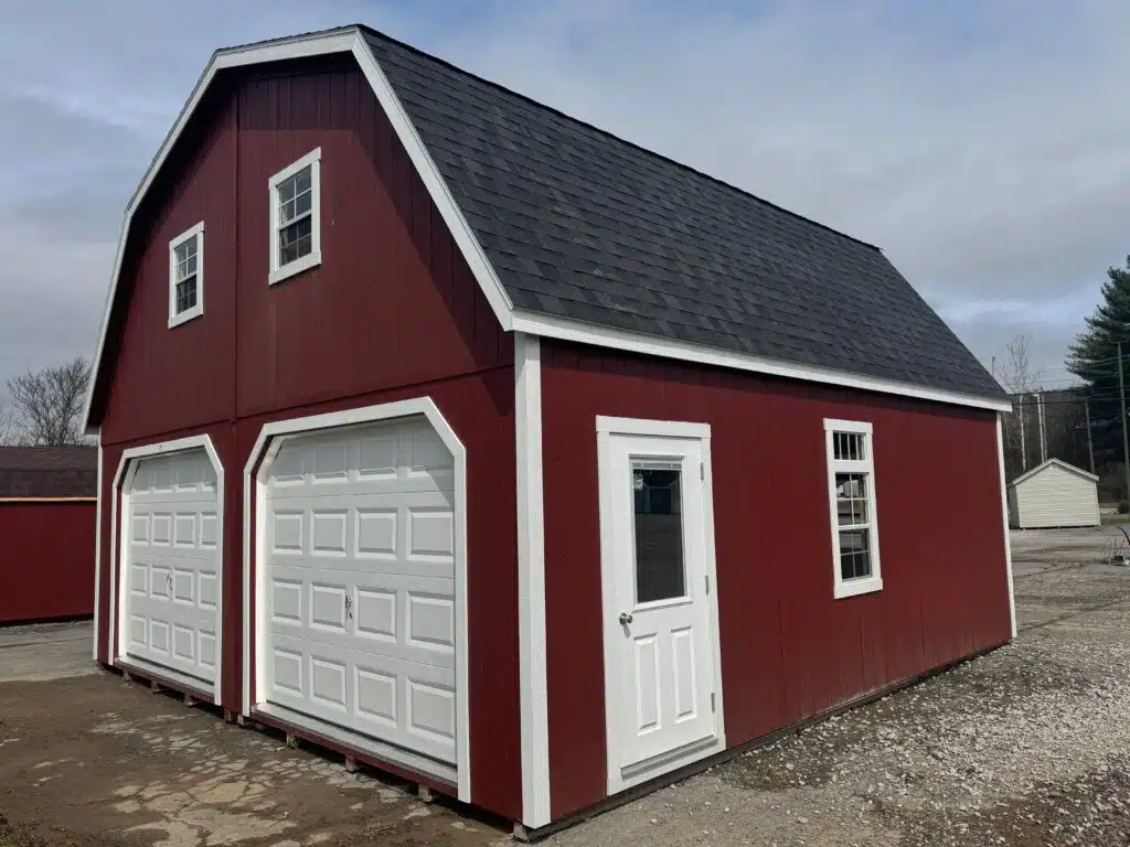 Red 24x24 two-story garage with white trim, black roof, two white 9x7 garage doors, white prehung half-lite entry door with blinds, upper windows, side window, and ridge vent shown from the front-side angle.